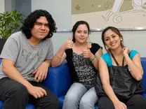 Tres personas sentadas en un sofá azul; la del centro levanta ambos pulgares.